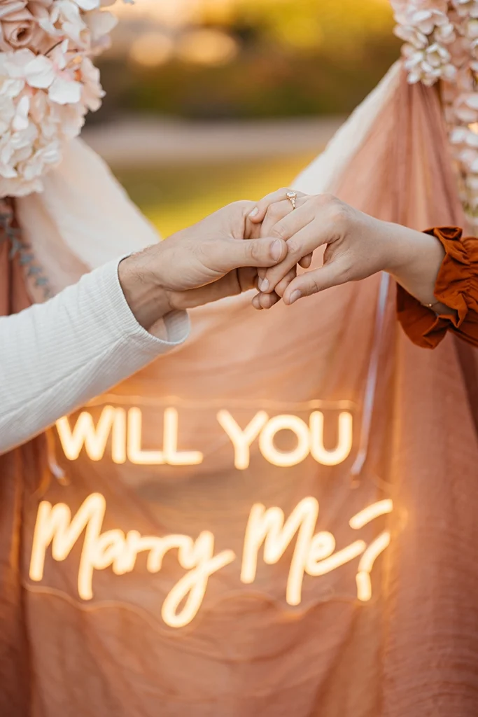 Detailaufnahme der Hände mit Verlobungsring vor einem leuchtenden Will You Marry Me Neonschild.