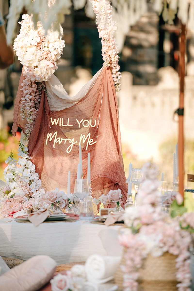 Detailaufnahme des leuchtenden Will You Marry Me Neonschilds auf drapiertem Stoff bei einem Heiratsantrag.