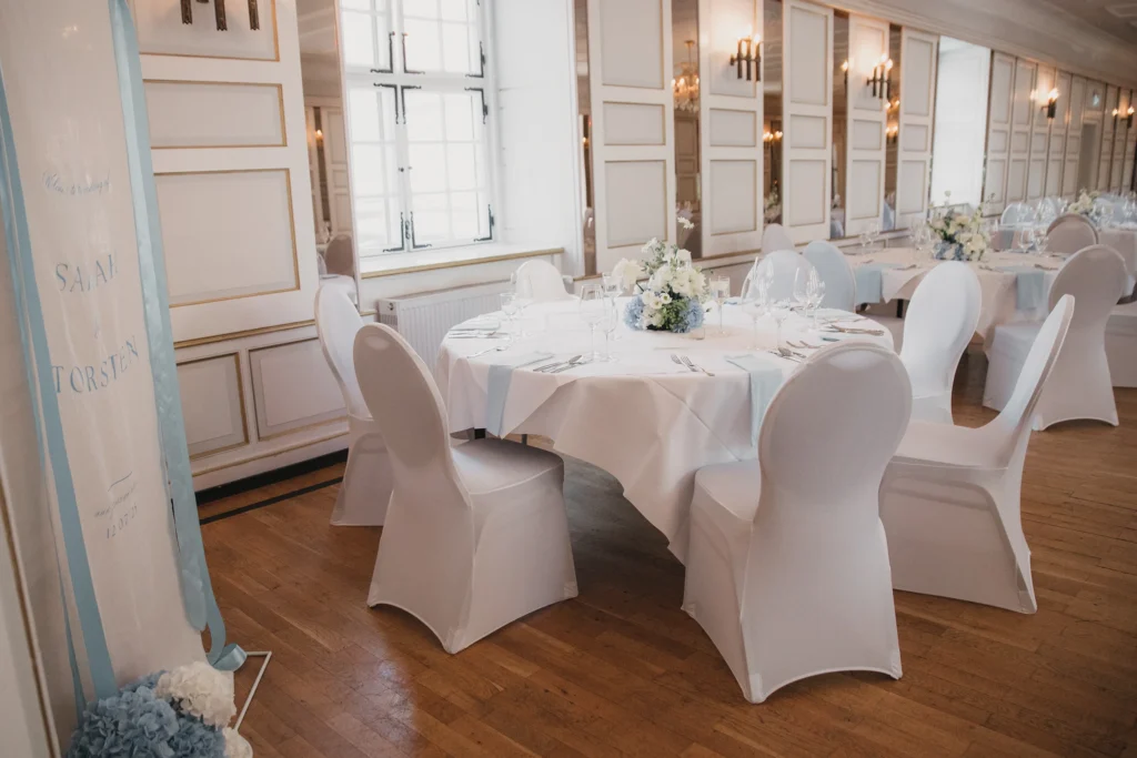 Rund gedeckte Hochzeitstafel mit weißen Stuhlhussen und Blumenschmuck im Haunsperger Saal