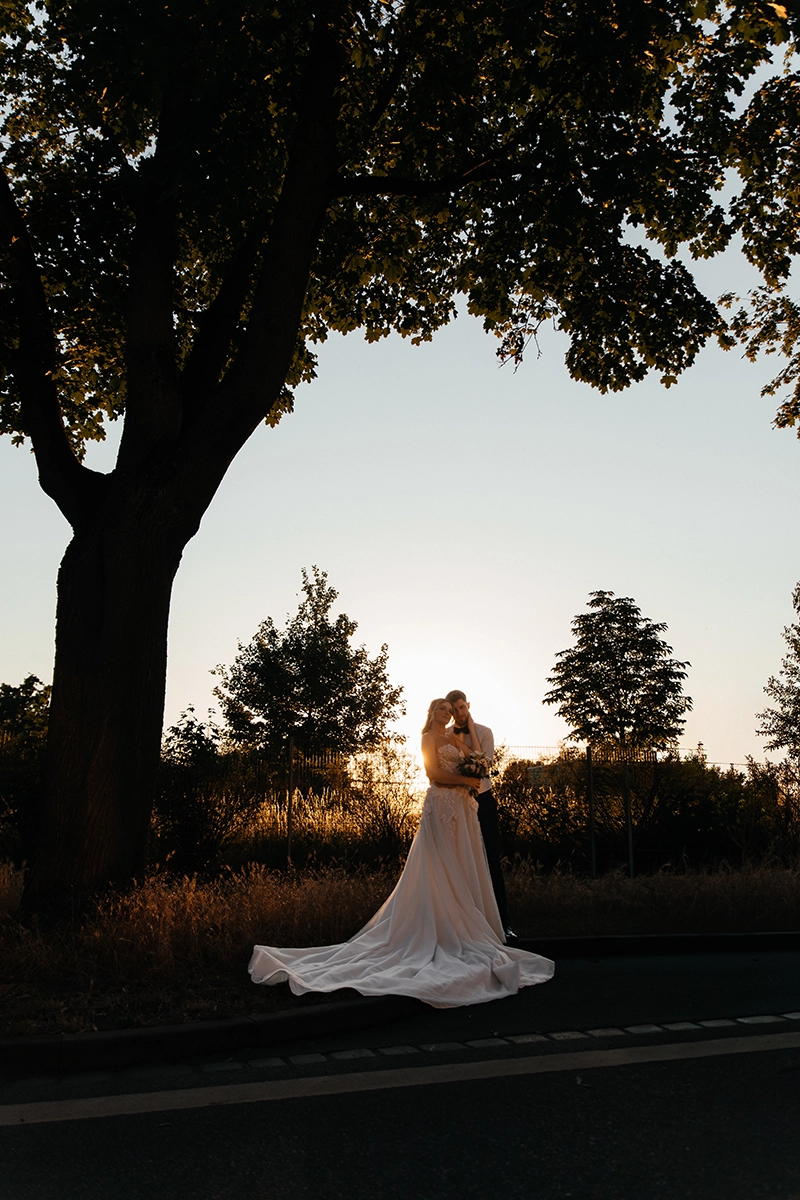Das Brautpaar steht mit ihrem langen Brautkleid und der ausgebreiteten Schleppe unter einem großen Baum im Abendlicht.
