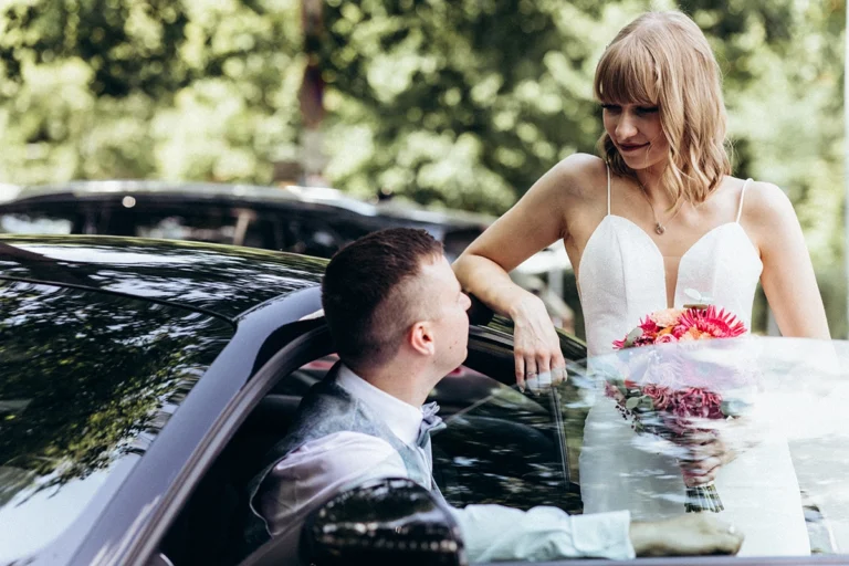 Die Braut steht mit ihrem Brautstrauß am geöffneten Hochzeitsauto und schaut den Bräutigam an.