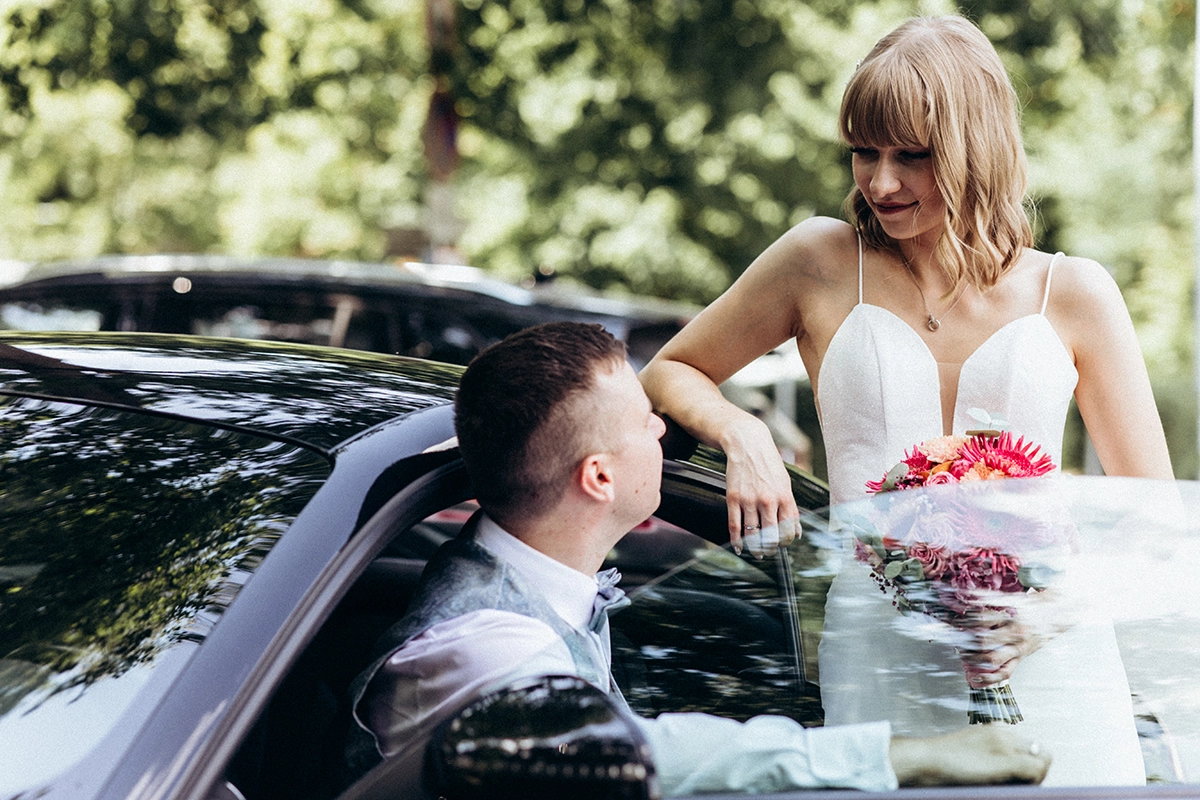 Die Braut steht mit ihrem Brautstrauß am geöffneten Hochzeitsauto und schaut den Bräutigam an.